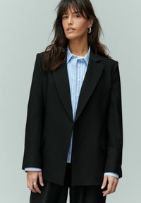 Blazer noir cintré avec un revers structuré et des poches latérales. Porté sur une chemise rayée bleu clair avec des détails aux poignets.