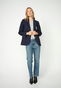 Tamsiai mėlynas švarkas su emblema kišenėje, dėvimas virš dryžuoto viršutinio drabužio ir derinamas su šviesiai mėlynais tiesiais džinsais bei juodomis smailiomis aukštakulnėmis.