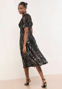 Rochie neagră din dantelă cu detalii sclipitoare argintii, cu mâneci scurte tip puff și tiv evazat. Purtați-o cu sandale negre cu curele, evidențiind o mișcare elegantă.