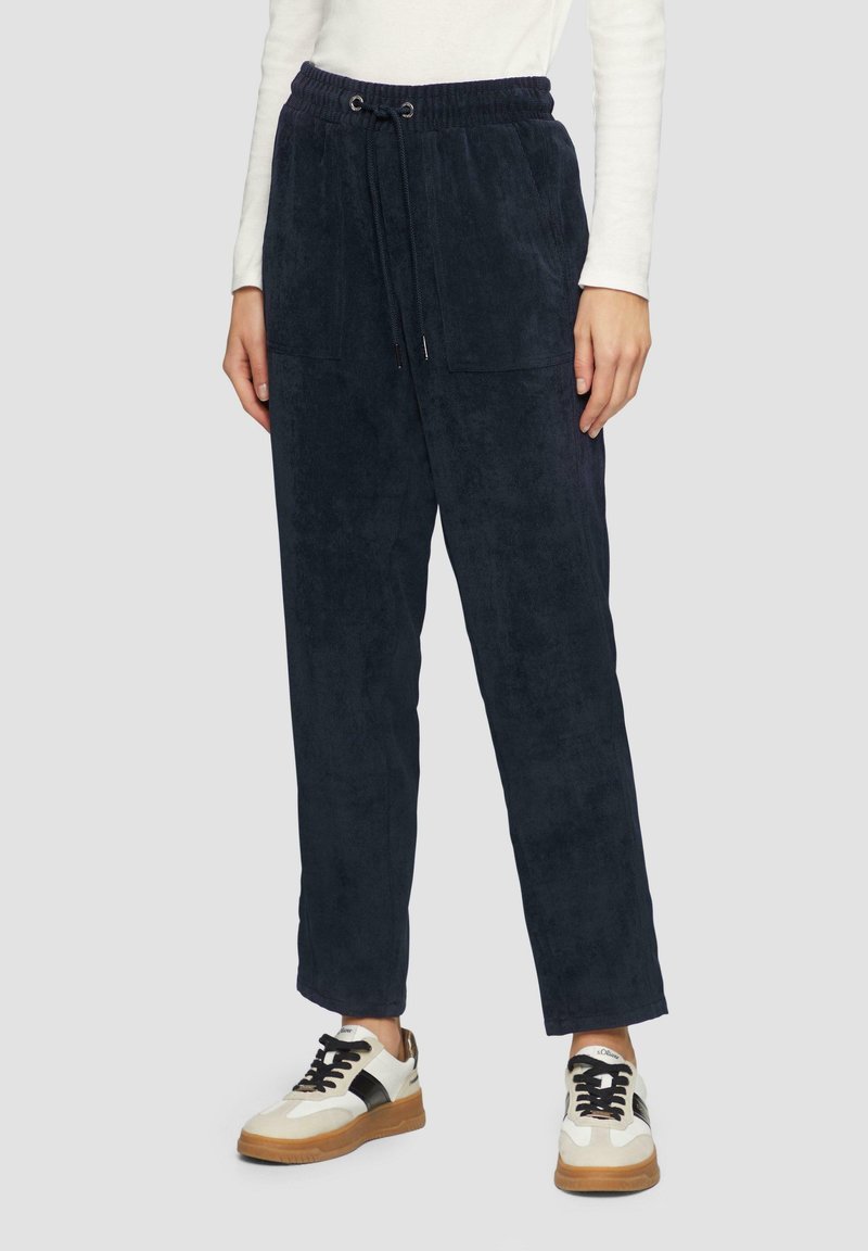 Personne portant un pantalon bleu foncé à cordon avec poches avant et des baskets blanches avec des accents noirs sur un fond clair.