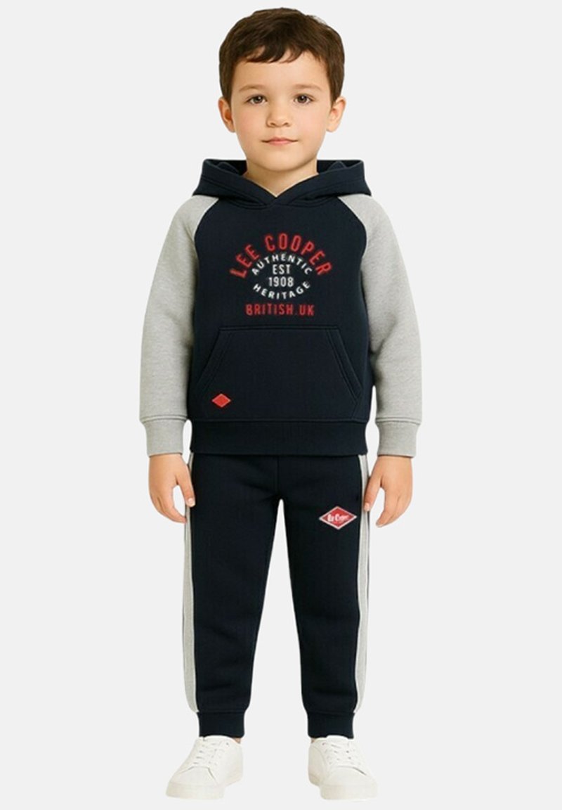 Sweat-shirt bleu marine avec manches grises et logo graphique rouge, associé à un jogging bleu marine avec des bandes latérales grises et un patch logo. Baskets blanches.