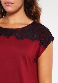 Kvinna som bär en vinröd topp med svart spetsdetaljering över halsringningen och axlarna, visar sin överkropp och läppar.