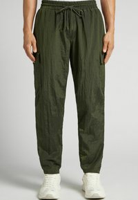 Groene, lichte cargobroek met een gekreukt textuur, elastische tailleband, trekkoord en zijzakken. Gecombineerd met witte sportschoenen.