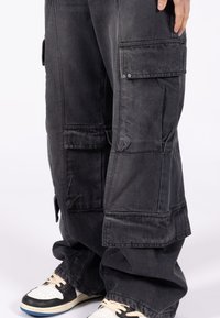 Osoba ubrana w luźne czarne spodnie cargo z wieloma kieszeniami oraz czarno-białe trampki z kremowymi sznurówkami na białym tle.