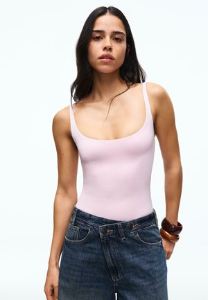 Vrouw met lang donker haar die een lichtroze tanktop, hooggetailleerde blauwe jeans en twee bruine armbanden aan haar rechterpols draagt.