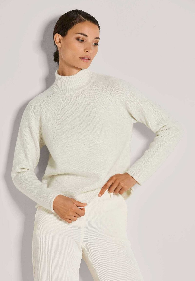 Weißer, gestrickter Pullover mit hochgeschlossenem, geripptem Kragen und Raglanärmeln, der eine weiche Textur und eine figurbetonte Silhouette aufweist, kombiniert mit passenden Hosen.