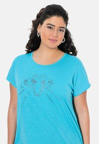 Camiseta turquoise de tela suave con un gráfico de tres caras de perro y la palabra "SELFIE" arriba en una impresión clara. Mangas cortas.