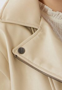 Beige neplederen jas met een geknikte kraag, zilveren rits en drukknoopdetails. Close-up afbeelding die de textuur en het ontwerp accentueert.