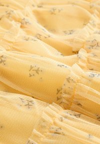 Geltono tiulio audinys su subtiliu gėlių raštu, kraštuose suklostytas raukiniais, minkštos tekstūros ir lengvas, orus dizainas.