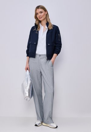 Młoda kobieta w granatowej kurtce, białej koszuli, szarych spodniach w prążki, srebrnych trampkach, trzymająca dużą srebrną metaliczną torbę, stoi na tle jednolitego jasnego tła.