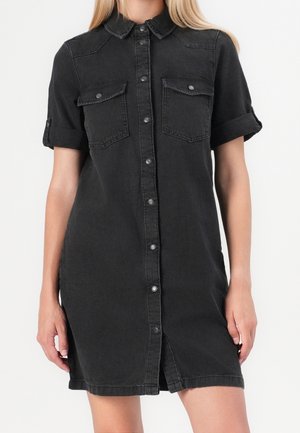 Kvinde iført en sort denimskjortekjole med korte ærmer, for- knapper og to brystlommer med klap foran mod en ensfarvet baggrund.