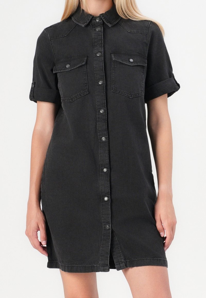 Femme portant une robe chemise en jean noire à manches courtes avec des boutons devant et deux poches poitrine à rabat sur un fond uni.
