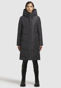 Pikk must puffer-jope kõrge kraega, vertikaalse tikandimustriga, tõmblukuga ja küljetaskutega, kantud mustade retuuside ja saapadega.