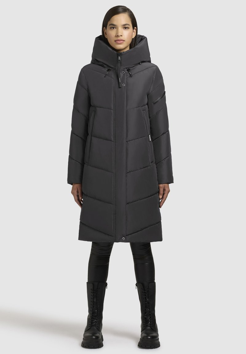 Pikk must puffer-jope kõrge kraega, vertikaalse tikandimustriga, tõmblukuga ja küljetaskutega, kantud mustade retuuside ja saapadega.