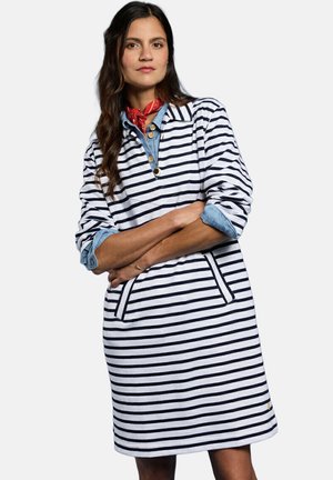 Femme aux longs cheveux foncés portant une robe rayée bleu marine et blanche sur une chemise bleue, avec un foulard rouge autour du cou, les bras croisés, debout devant un fond uni.