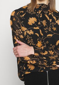 Blouse noire avec un motif floral dans des tons orange, en tissu transparent, col haut, manches longues avec poignets élastiques, et silhouette décontractée.