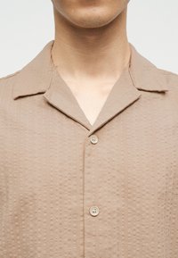 Άτομο που φοράει μπεζ υφασμάτινο κοντομάνικο πουκάμισο με γιακά τύπου κάμπινγκ και δύο ορατά κουμπιά, εμφανιζόμενο από τον λαιμό μέχρι το μέσο του στήθους.