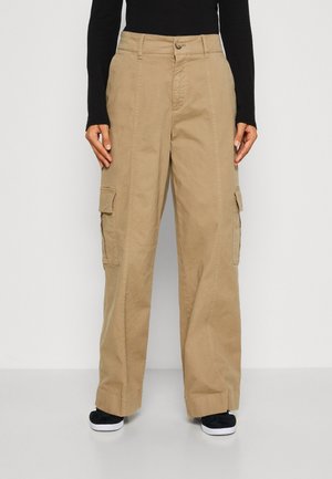 Khakiowe spodnie cargo o szerokich nogawkach z bocznymi kieszeniami na klapy, noszone przez osobę w czarnym długim rękawie i czarnych tenisówkach z białymi podeszwami.