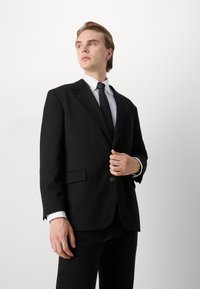 Melnā pielāgotā žakete ar ieplestiem atlociem un vienu pogu, saskaņota ar baltu kreklu un melnu kaklasaiti, uzvilkta virs tumšiem džinsiem.