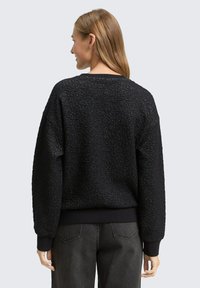 Schwarzes Sweatshirt mit strukturiertem Material, lockerem Schnitt, Rippbündchen und -saum. Das gestrickte Muster verleiht Tiefe und steht im Kontrast zum glatten Stoff.