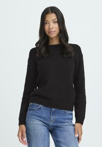 Ung kvinde med langt sort hår iført en struktureret sort sweater og blå jeans, stående foran en ensfarvet lys baggrund.