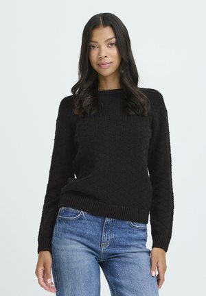 Young woman with long black hair wearing a textured black sweater and blue jeans, standing against a plain light background.