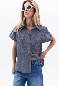 Svart- och vitrutigt kortärmat skjortor med knapad front, tillsammans med ljusblå högmidjade jeans. Modellen har på sig vita solglasögon.