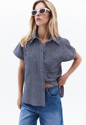 Chemise à carreaux noir et blanc à manches courtes avec un bouton sur le devant, associée à un jean taille haute bleu clair. Le modèle porte des lunettes de soleil blanches.