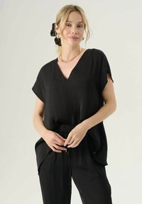 Čierna top s výstrihom do V vyrobený z ľahkého materiálu, s krátkymi rukávmi a voľným strihom. Skombinovaný s čiernymi nohavicami na sťahovanie a zlatými šperkovými akcentmi.