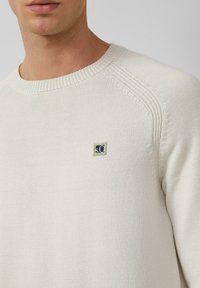 Man in een cr�èmekleurige gebreide trui met een geribde kraag en een klein rechthoekig logo-embleem op de borst.