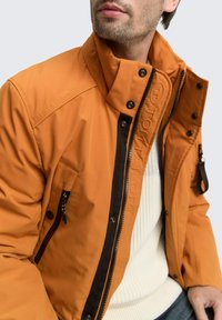 Oranje waterdichte jas met hoge kraag, zwarte rits en meerdere zakken. Beschikt over een gestructureerde stof en een verhoogd merkembleem aan de voorkant.