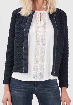 Lyshåret kvinde iført en tekstureret sort quiltet jakke over en hvid bluse med blondekant og blå jeans, stående med afslappede hænder.