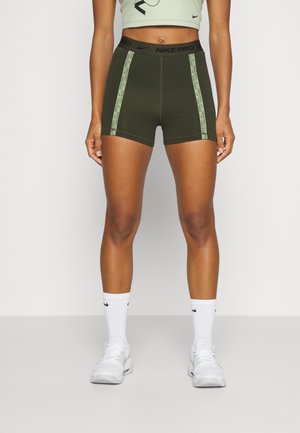 Frau trägt olivgrüne Nike Pro Sportshorts mit gebrandeten Seitenstreifen, weiße Nike-Socken und weiße Sneakers, steht vor einem schlichten Hintergrund.