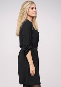 Blonde vrouw in profiel, gekleed in een zwart, met een riem accentueerd jurkje met opgerolde mouwen, staande tegen een effen lichte achtergrond.