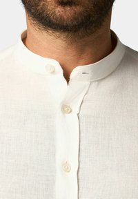 Camicia bianca in lino con colletto alla mandarina, caratterizzata da una pattina con due bottoni e un leggero motivo testurizzato.
