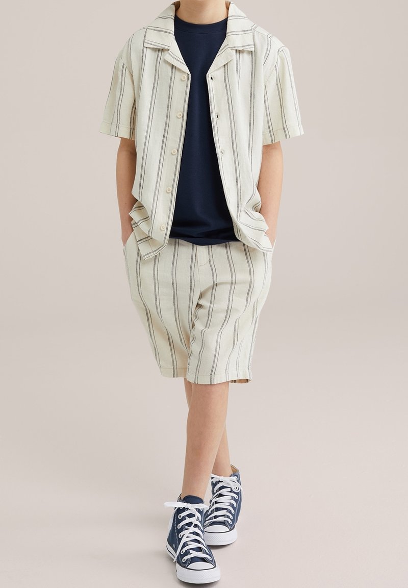 Enfant portant une chemise à manches courtes à rayures verticales crème et un short, avec un t-shirt bleu marine et des baskets montantes bleu marine, debout.