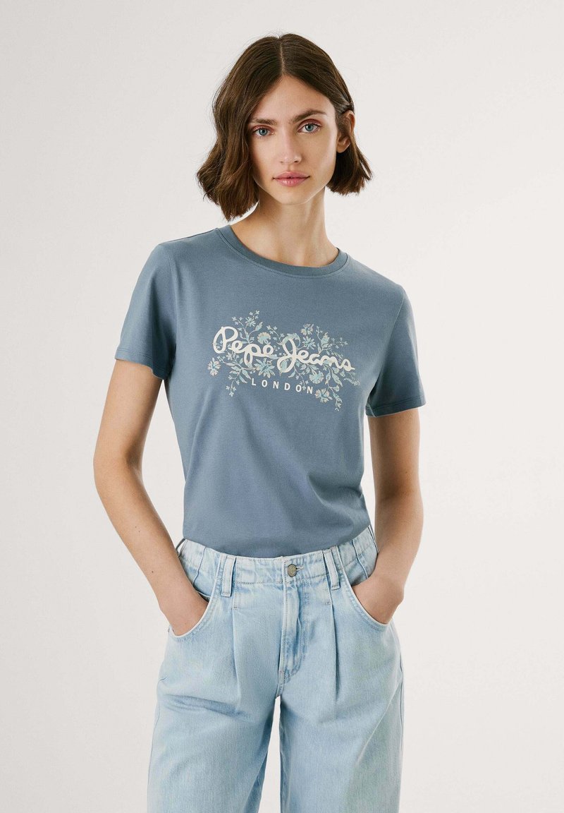 Zils kokvilnas T-krekls ar baltu ziedu grafiku un tekstu, apvienots ar gaišiem džinsu biksēm ar krokām un sānu kabatām.