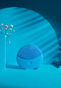 Sirkulær blå ansiktsrensingsenhet med teksturert silikonoverflate, aksentert med knapper, satt mot en lys blå bakgrunn med blomster.