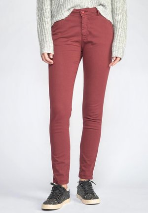 Person wearing fitted dark red pants, gray knit sweater, and black speckled sneakers, standing against a light gray background.