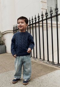 Marineblauwe kabelgebreide trui, losse verwassen spijkerbroek, marineblauwe schoenen met witte veters, staand op een stenen oppervlak bij een zwart ijzeren hek.