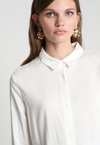 Blusa bianca a maniche lunghe con chiusura a bottoni e una texture morbida e liscia. Presenta un colletto classico e una leggera lucentezza. Orecchini rotondi trasparenti.