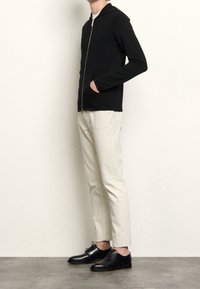 Svart zip-up hoodie med framficka. Matchas med krämfärgade, slim-fit byxor och blanka svarta dresskängor. Enkel, modern design.