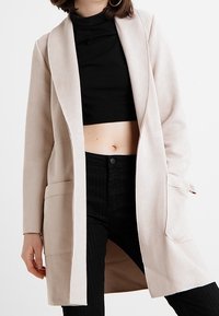 Donna che indossa un lungo cappotto beige aperto su un dolcevita nero corto e pantaloni neri a vita alta, in piedi contro uno sfondo bianco.