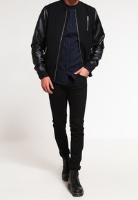 Chaqueta bomber negra con mangas de cuero, cierre delantero y bolsillos laterales. Llevada sobre una camisa oscura de botones y pantalones negros, combinada con botas.