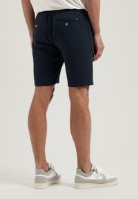 Pantaloni scurți bleumarin dintr-un material texturat, cu talie elastică și două buzunare pe spate cu închidere prin buton. Modelul este asociat cu adidași gri și albaștri.