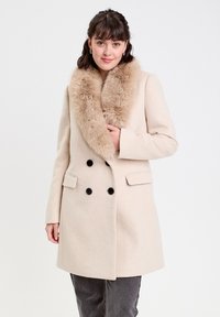 Manteau en laine beige avec un col en fausse fourrure, design croisé avec des boutons noirs et des poches latérales, présenté sur un fond neutre.