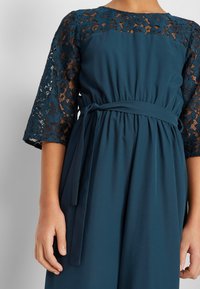 Femme portant une robe teal avec des manches en dentelle mi-longues et une ceinture nouée à la taille, les mains détendues sur les côtés devant un fond uni.