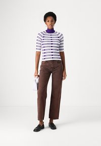 Top a maniche corte a righe viola e bianche con scollatura alta, jeans dritti marroni, scarpe nere e una piccola borsa bianca.