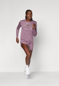 Woman running in light purple long-sleeve shirt and shorts with white sneakers against plain white background.