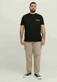 Black cotton t-shirt with a front pocket detail, paired with beige pants and black sneakers. Man standing against a neutral background.